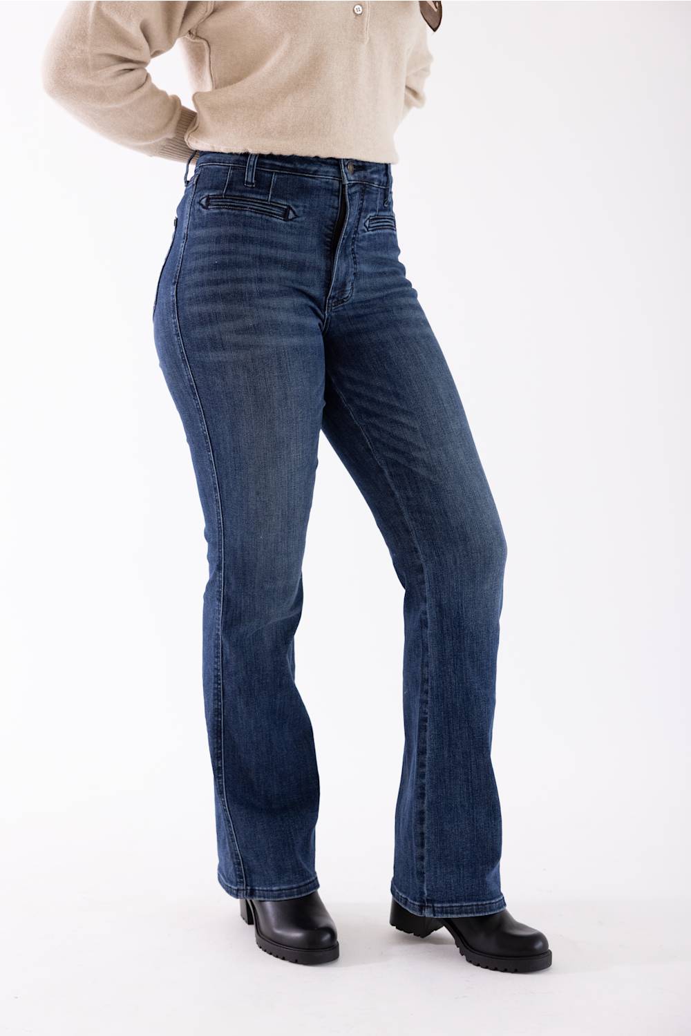 A person wearing dark blue high-rise flare jeans, a beige long-sleeve top, and black leather boots, standing against a plain white studio background.