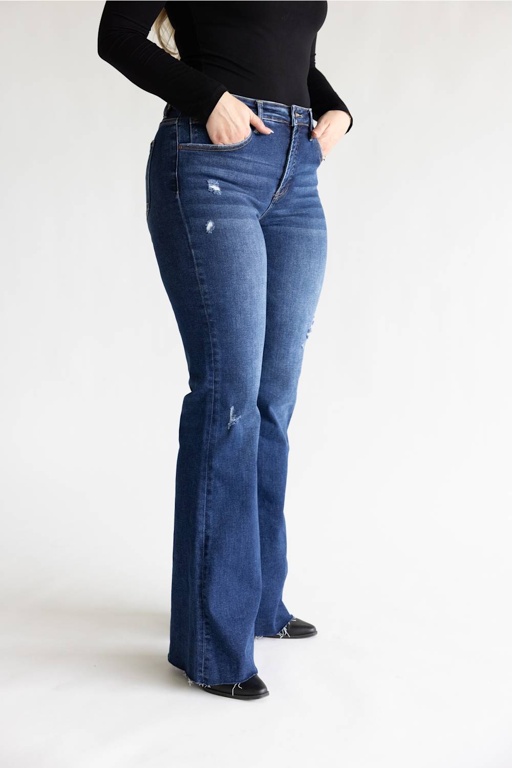 A person wearing dark blue high-waisted flare jeans with distressed details, paired with a black long-sleeve top and black shoes, standing against a plain white studio background.