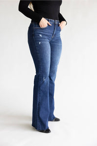 A person wearing dark blue high-waisted flare jeans with distressed details, paired with a black long-sleeve top and black shoes, standing against a plain white studio background.