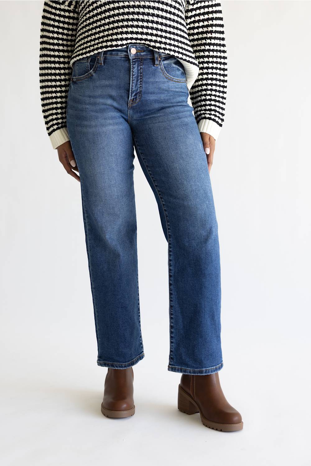 A person wearing medium-wash blue straight-leg jeans, a black and white houndstooth sweater, and brown ankle boots, standing against a plain white studio background.