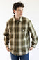 A man wearing a Carhartt long-sleeve plaid button-up shirt in olive green, brown, and beige, paired with blue jeans, against a plain white background.
