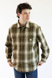 A man wearing a Carhartt long-sleeve plaid button-up shirt in olive green, brown, and beige, paired with blue jeans, against a plain white background.