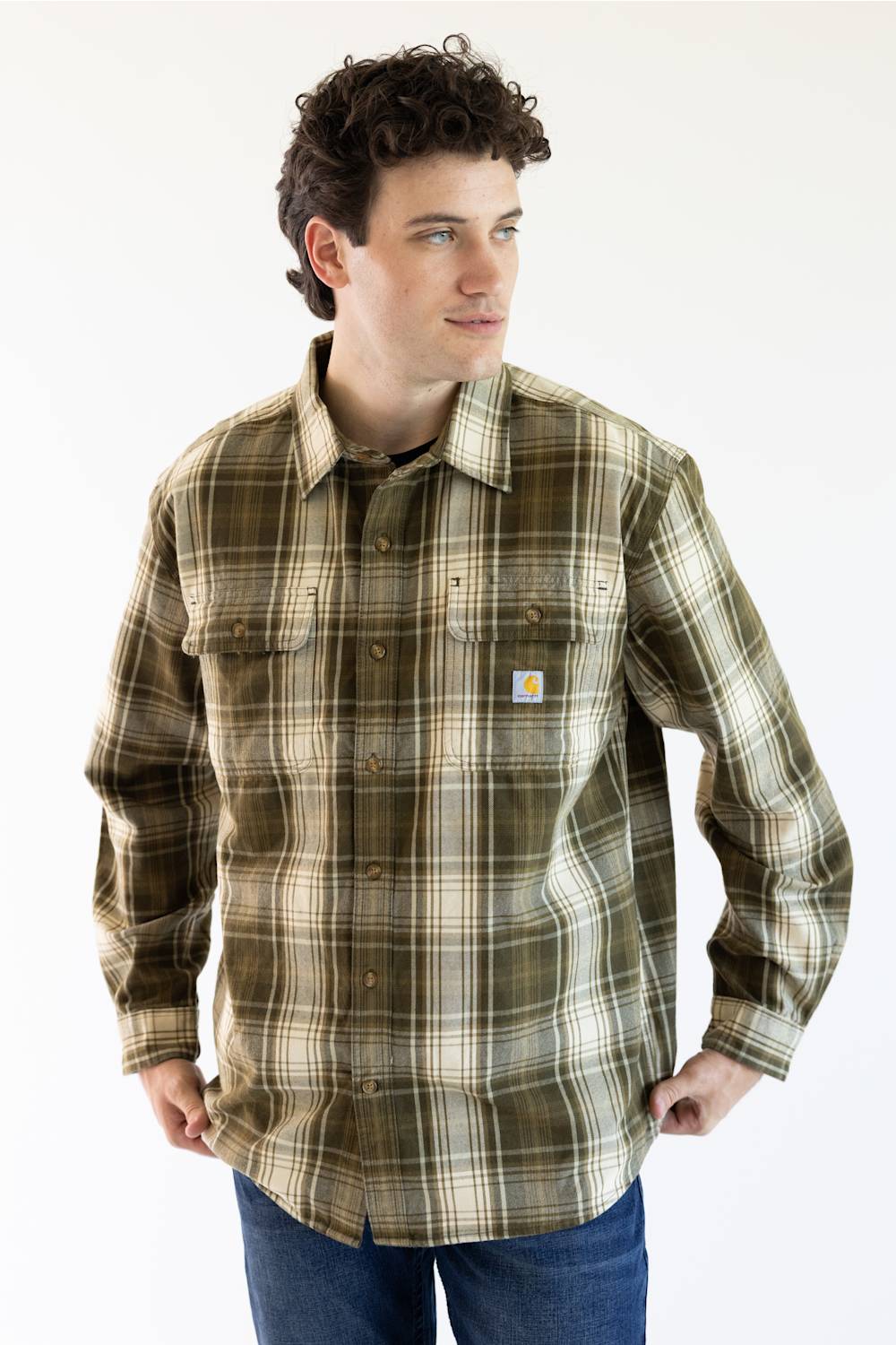 A man wearing a Carhartt long-sleeve plaid button-up shirt in olive green, brown, and beige, paired with blue jeans, against a plain white background.