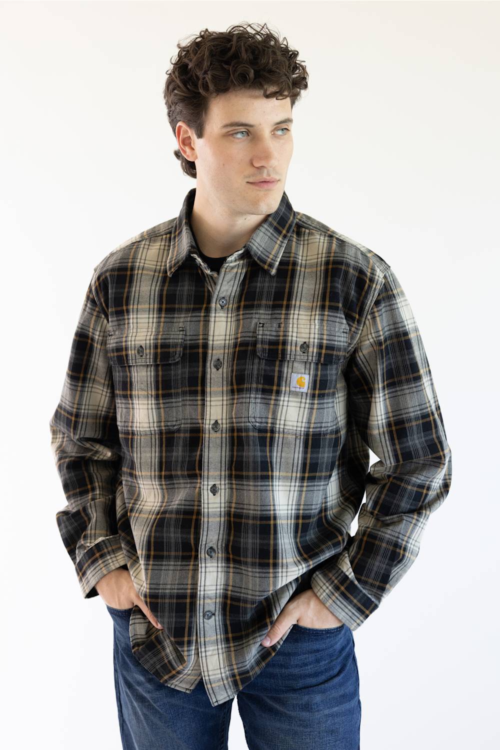 A man wearing a Carhartt plaid button-down shirt in black, gray, and tan, paired with blue jeans, standing against a plain white background.