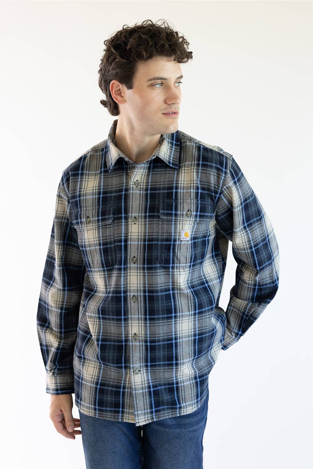 A man wearing a Carhartt long-sleeve plaid button-down shirt in navy, blue, and beige, paired with blue jeans, photographed against a plain white studio background.