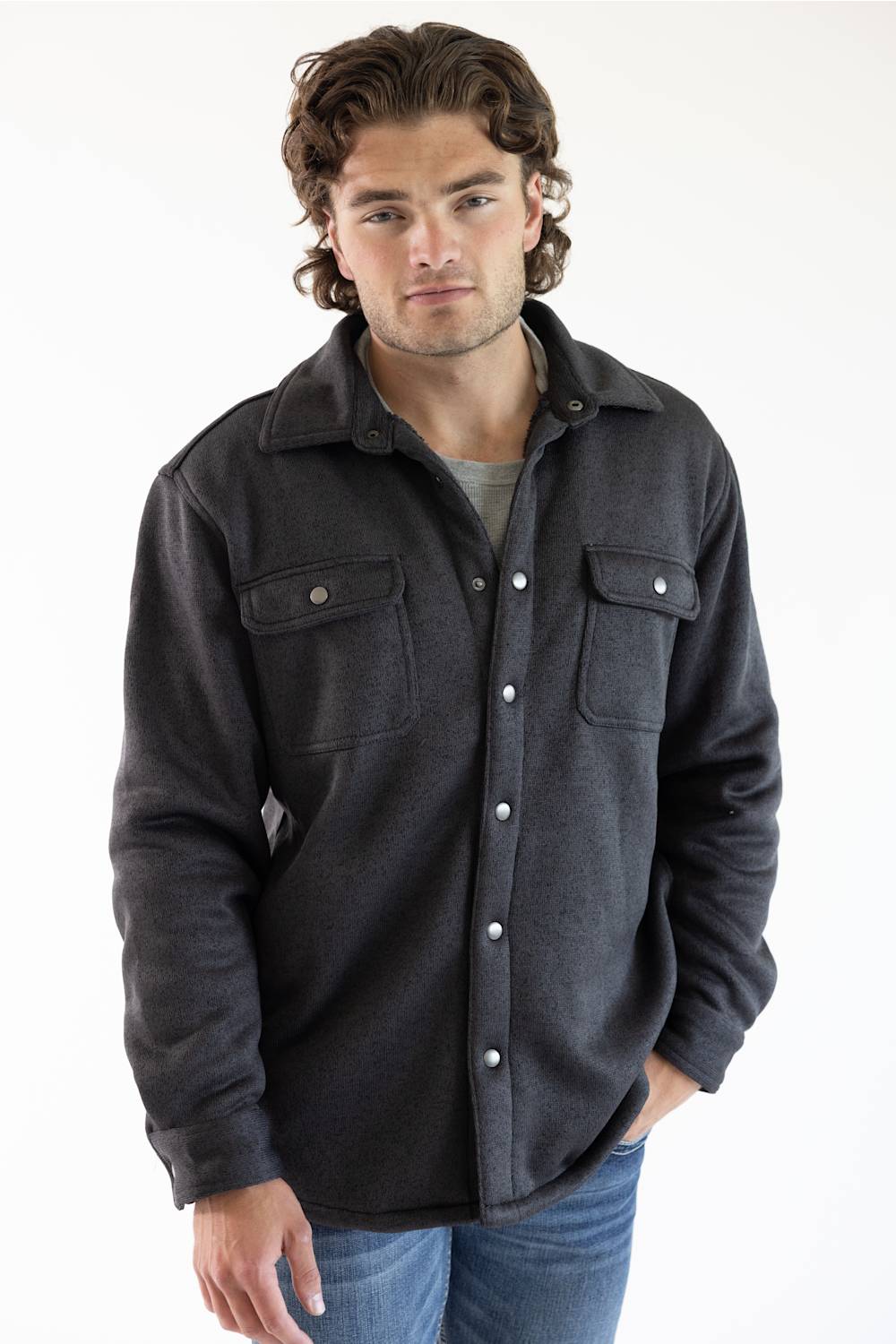 A man wearing a dark charcoal gray button-up flannel shirt jacket with two chest pockets and silver snap buttons, paired with blue jeans.