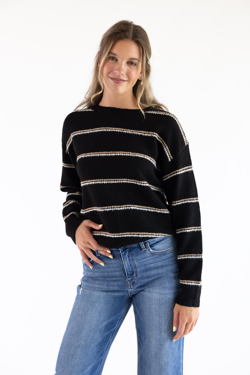 Smiling model styled in a black crew neck knit sweater with textured tan and white embroidered horizontal stripes, paired with high-waisted blue jeans against a plain white background.