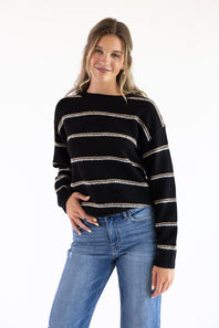 Smiling model styled in a black crew neck knit sweater with textured tan and white embroidered horizontal stripes, paired with high-waisted blue jeans against a plain white background.