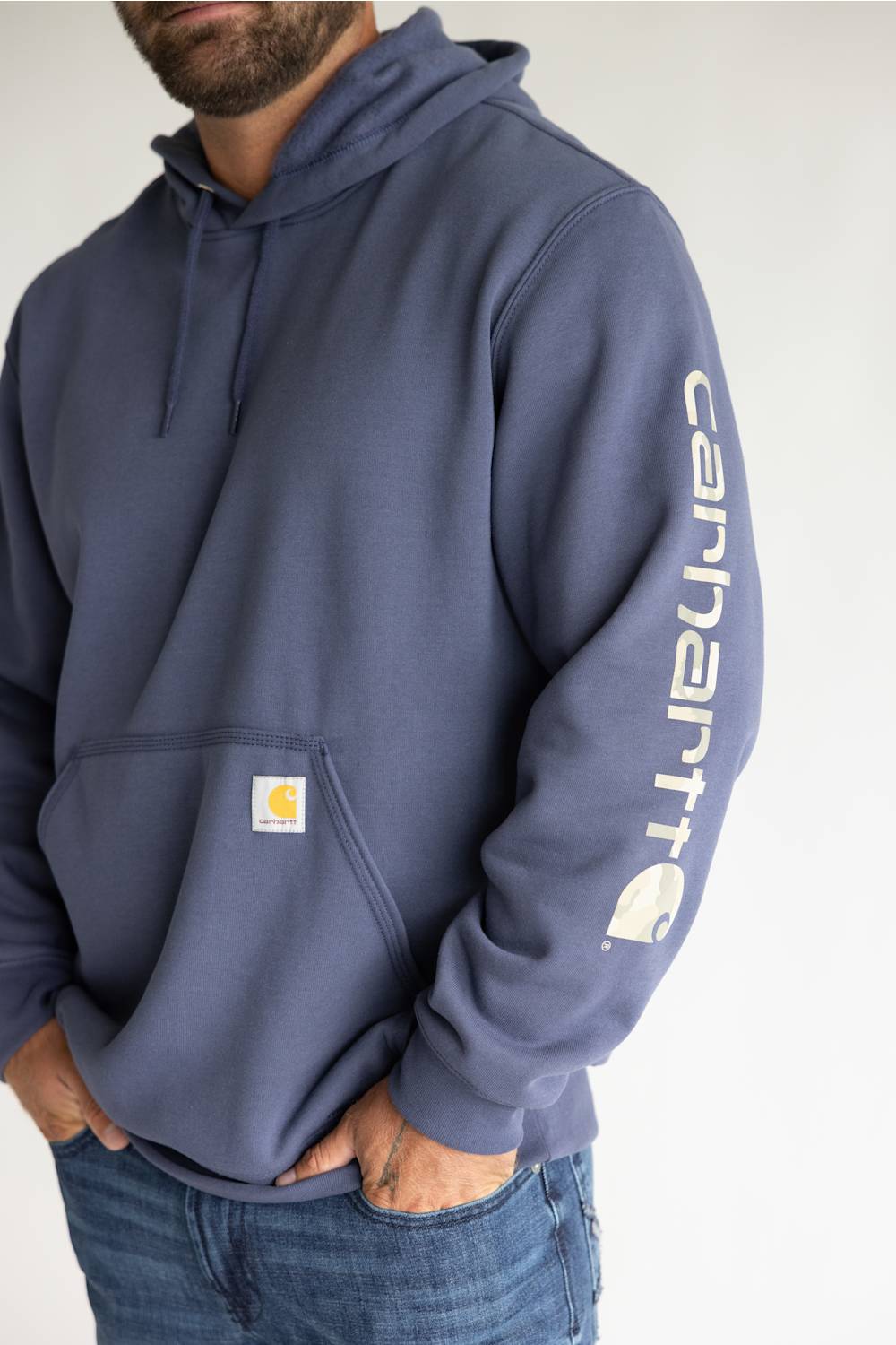 A man wearing a navy blue Carhartt pullover hoodie with a white vertical Carhartt logo printed on the left sleeve and a small signature yellow-and-white Carhartt patch on the front pocket, paired with blue jeans against a plain white background.