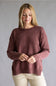 A woman wearing a dusty rose knit crewneck sweater and light beige pants, standing against a neutral gray studio background.