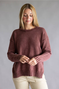 A woman wearing a dusty rose knit crewneck sweater and light beige pants, standing against a neutral gray studio background.