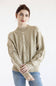 A woman wearing a beige cable-knit sweater with cream-colored braided patterns on the front and sleeves, paired with blue jeans, against a plain white background.
