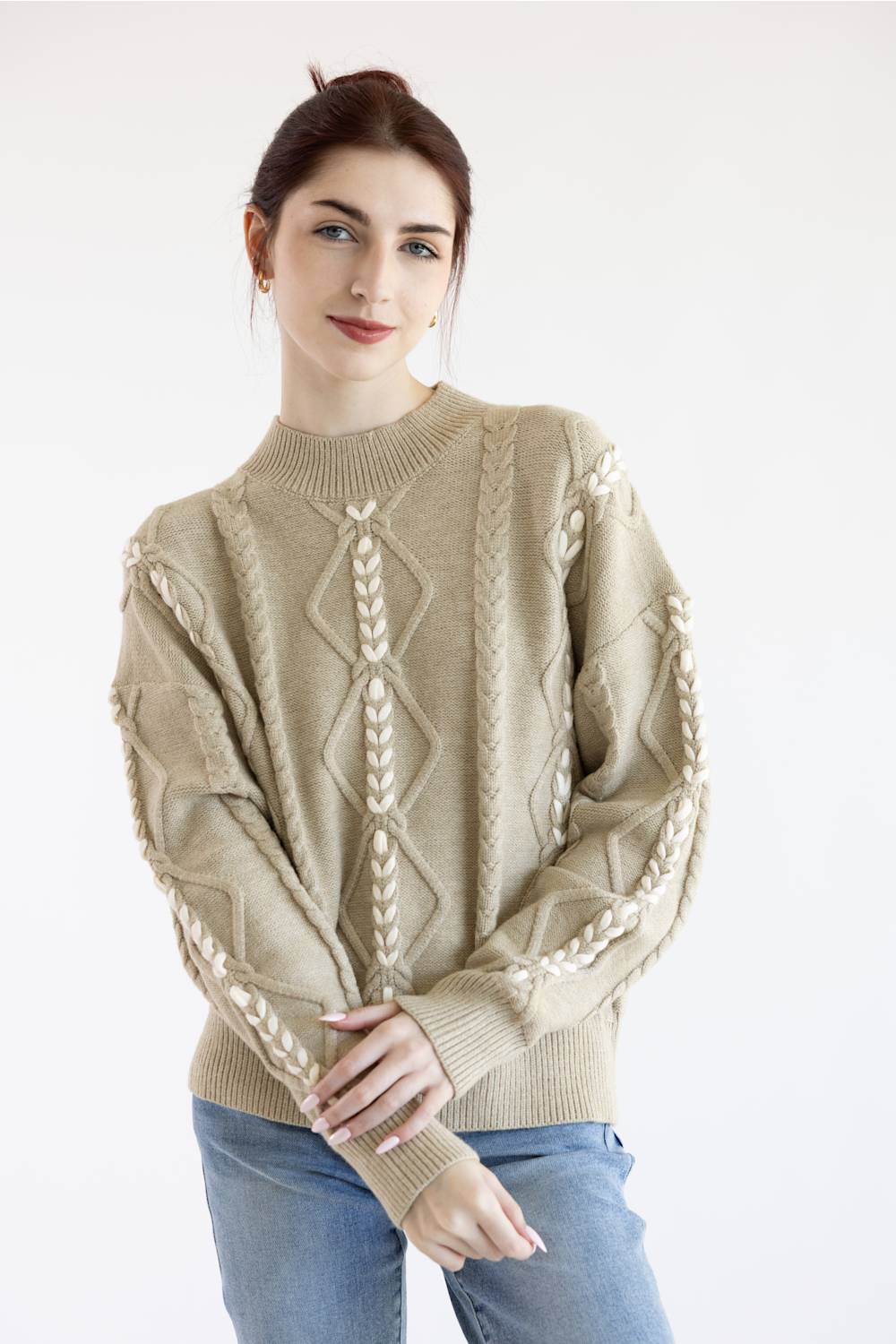 A woman wearing a beige cable-knit sweater with cream-colored braided patterns on the front and sleeves, paired with blue jeans, against a plain white background.