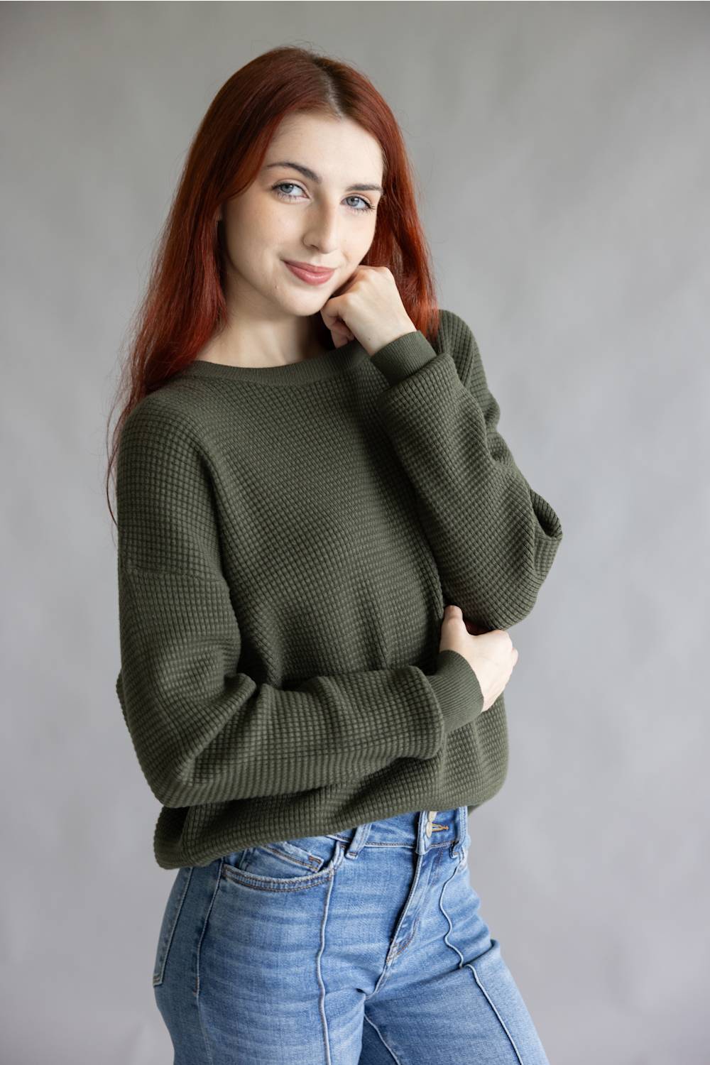 A woman with red hair wearing an olive green waffle-knit sweater and blue jeans, posing against a neutral gray studio background.