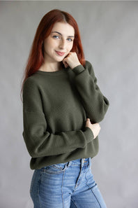 A woman with red hair wearing an olive green waffle-knit sweater and blue jeans, posing against a neutral gray studio background.