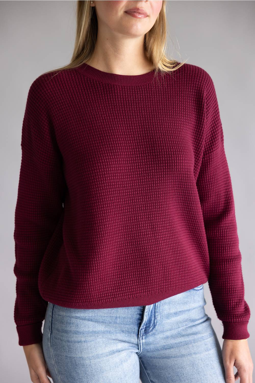 A woman wearing a long-sleeve waffle-knit crewneck sweater in deep burgundy, paired with light blue jeans, against a plain gray studio background.