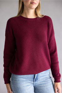 A woman wearing a long-sleeve waffle-knit crewneck sweater in deep burgundy, paired with light blue jeans, against a plain gray studio background.