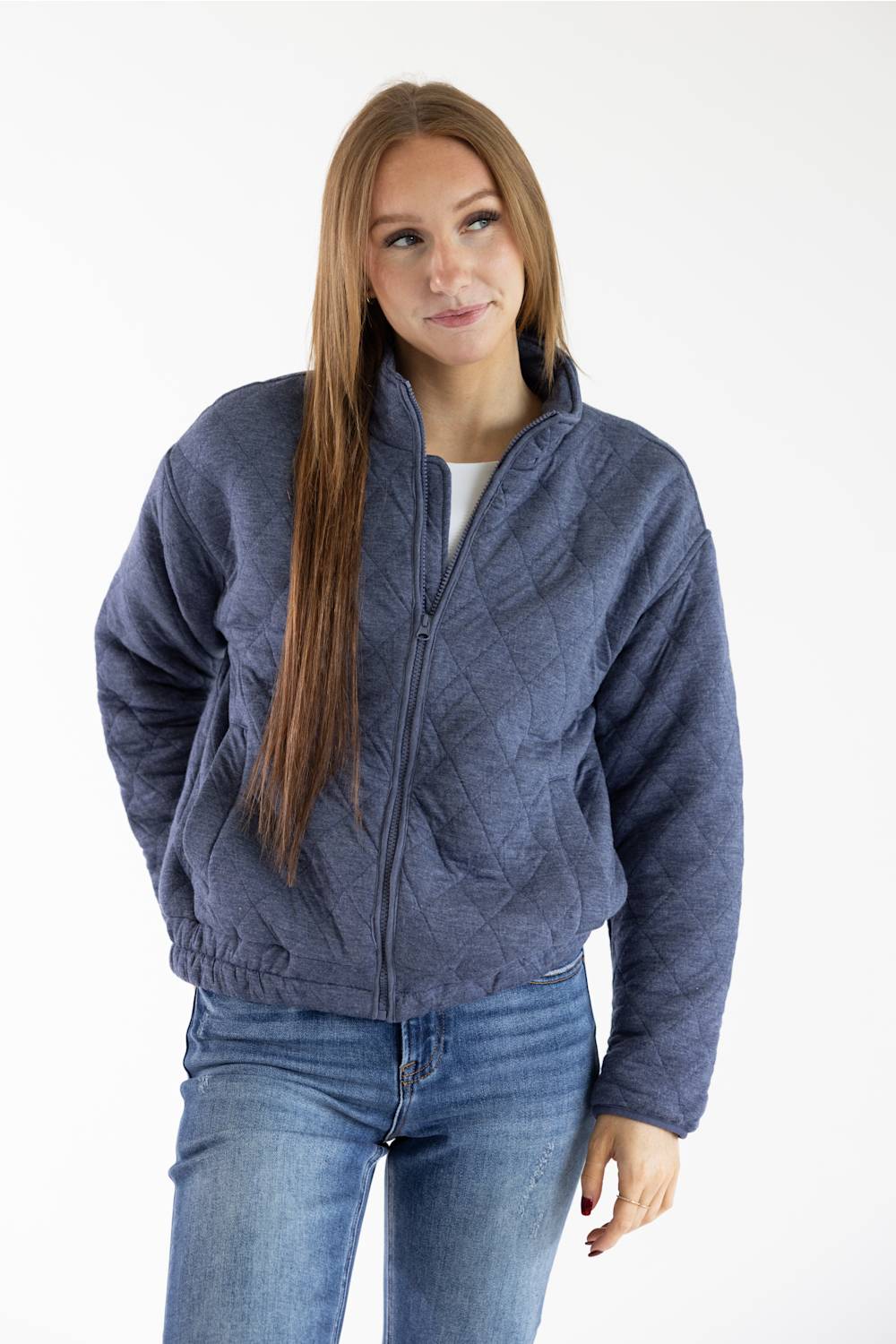 A woman modeling a slate blue quilted zip-up jacket with a stand collar, paired with blue jeans, against a plain white studio background.