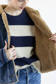 A close-up of a person wearing a navy and cream striped knit sweater under an open brown sherpa-lined denim jacket, paired with blue jeans against a plain white background.