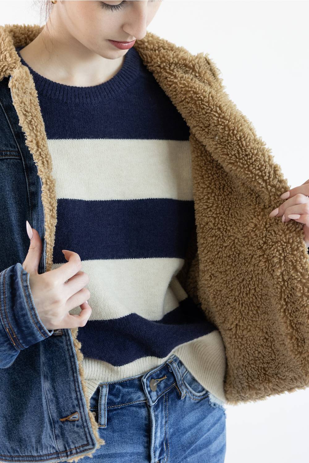 A close-up of a person wearing a navy and cream striped knit sweater under an open brown sherpa-lined denim jacket, paired with blue jeans against a plain white background.
