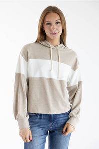 A woman wearing a color-blocked knit hoodie with beige, white, and taupe horizontal stripes, paired with blue jeans, against a plain white studio background.