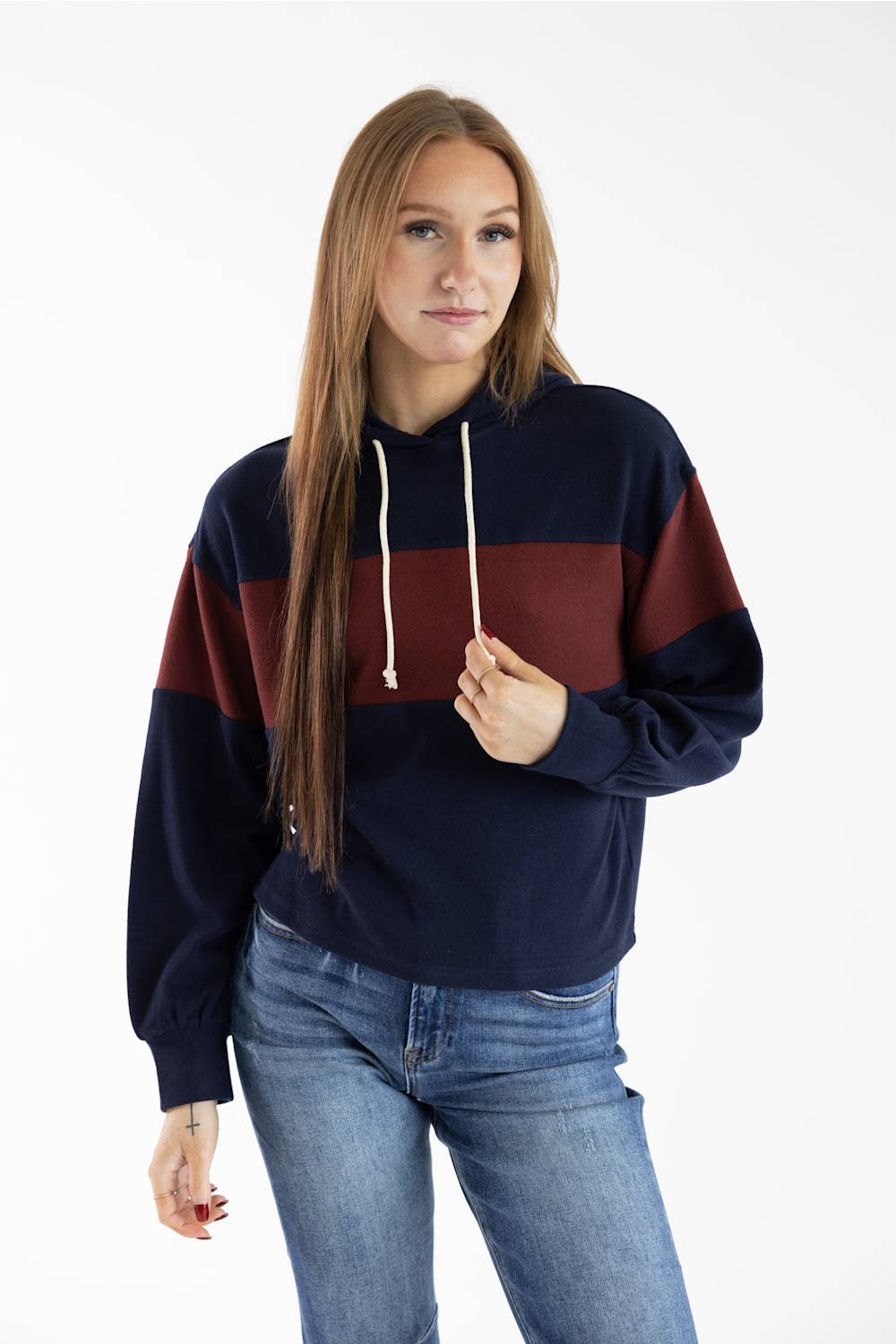 A woman modeling a color-blocked pullover hoodie with navy blue, burgundy, and dark gray horizontal stripes, paired with blue jeans, against a plain white studio background.