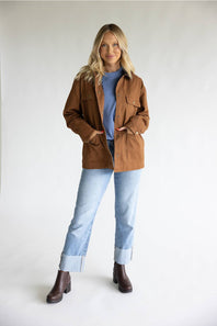 A woman modeling a brown utility jacket over a blue t-shirt, light-wash jeans, and brown leather ankle boots, standing against a plain white studio background.