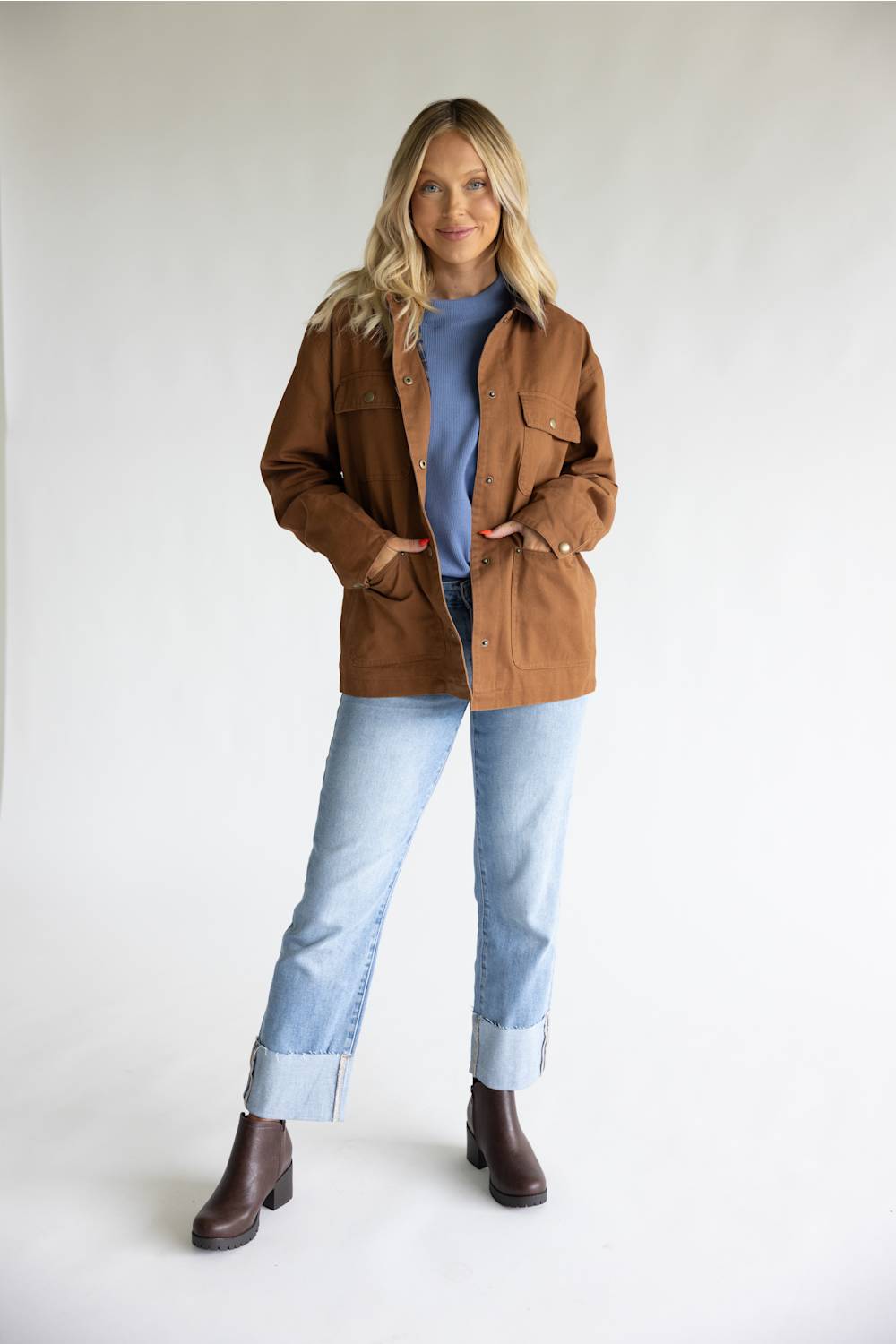 A woman modeling a brown utility jacket over a blue t-shirt, light-wash jeans, and brown leather ankle boots, standing against a plain white studio background.