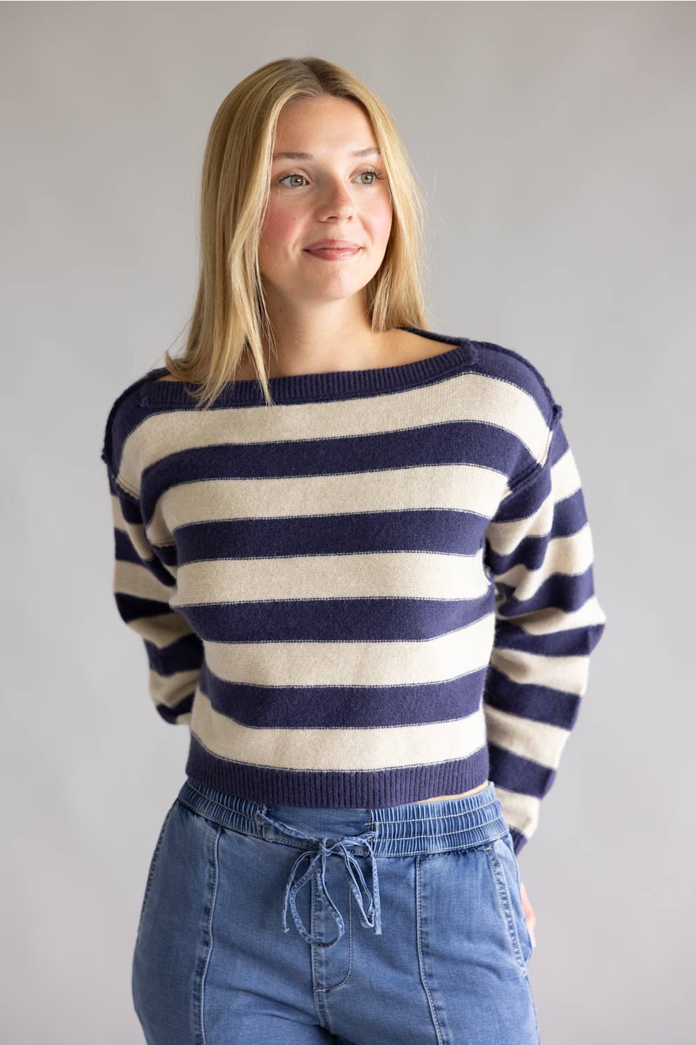 A woman wearing a navy blue and cream striped knit sweater with blue jeans, standing against a plain light gray studio background.
