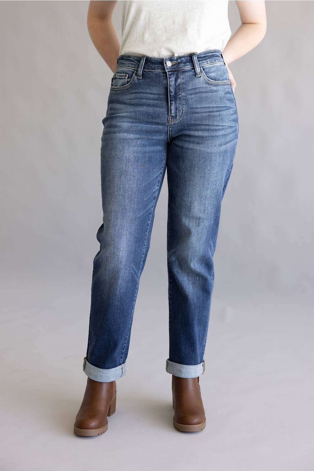 A person wearing medium-wash blue denim jeans with rolled cuffs and brown ankle boots, standing against a plain light gray studio background.
