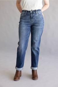 A person wearing medium-wash blue denim jeans with rolled cuffs and brown ankle boots, standing against a plain light gray studio background.