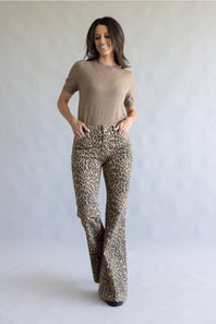 A woman modeling leopard-print flared pants and a beige short-sleeve top, standing against a light gray studio backdrop.