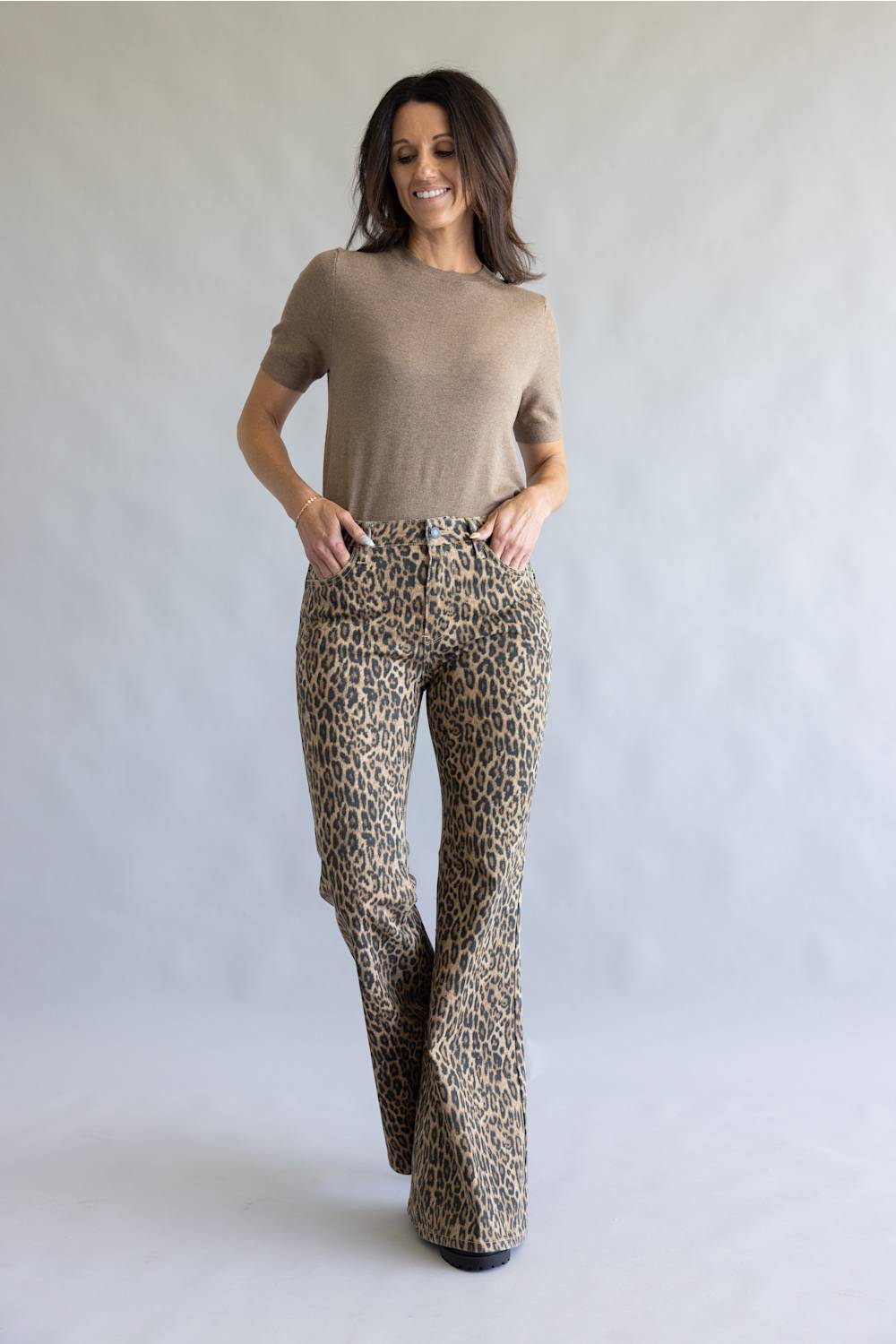 A woman modeling leopard-print flared pants and a beige short-sleeve top, standing against a light gray studio backdrop.