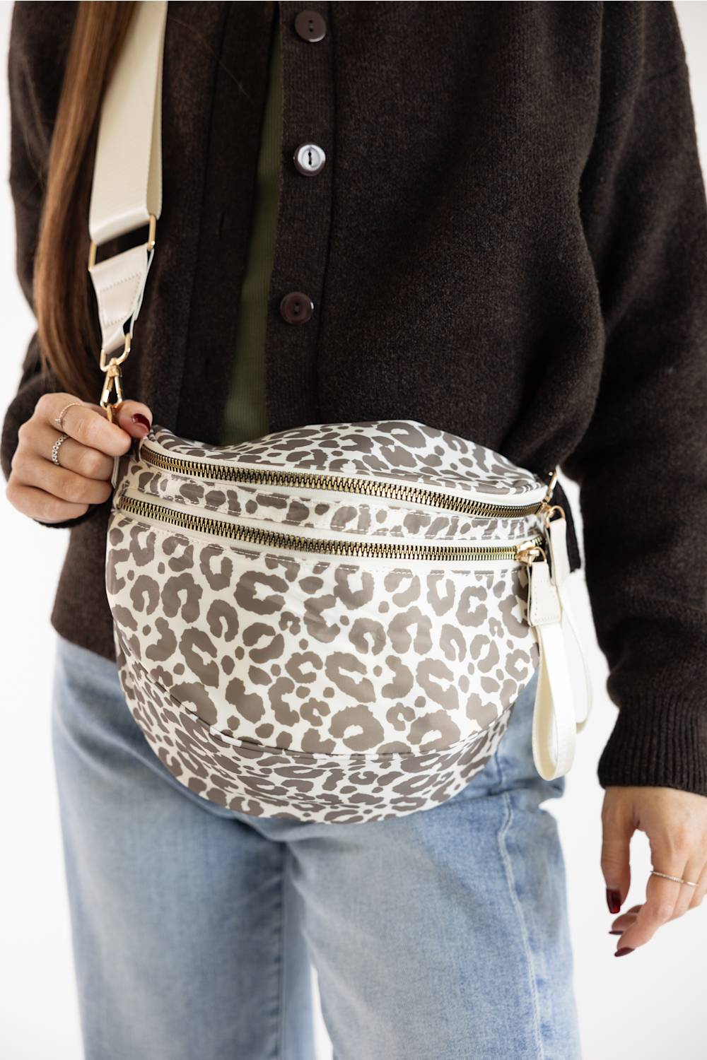 A person wearing a brown knit cardigan and light blue jeans carries a leopard-print crossbody bag with a beige strap and gold-tone zipper hardware.