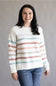 A woman smiling while wearing a cream knit sweater with horizontal stripes in dusty pink, light blue, and gray, paired with blue jeans, against a plain gray studio background.