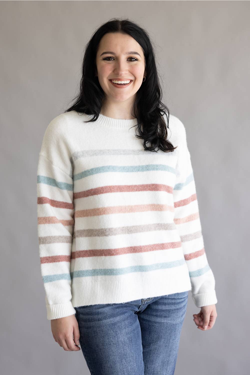 A woman smiling while wearing a cream knit sweater with horizontal stripes in dusty pink, light blue, and gray, paired with blue jeans, against a plain gray studio background.