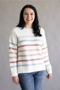 A woman smiling while wearing a cream knit sweater with horizontal stripes in dusty pink, light blue, and gray, paired with blue jeans, against a plain gray studio background.