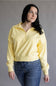 A woman wearing a pale yellow half-zip sweatshirt and blue jeans, standing against a plain gray studio background.