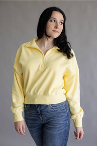 A woman wearing a pale yellow half-zip sweatshirt and blue jeans, standing against a plain gray studio background.