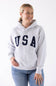 Front view of a blonde woman wearing an ice grey Reflex fleece pullover hoodie with black "USA" block lettering across the chest and blue jeans.