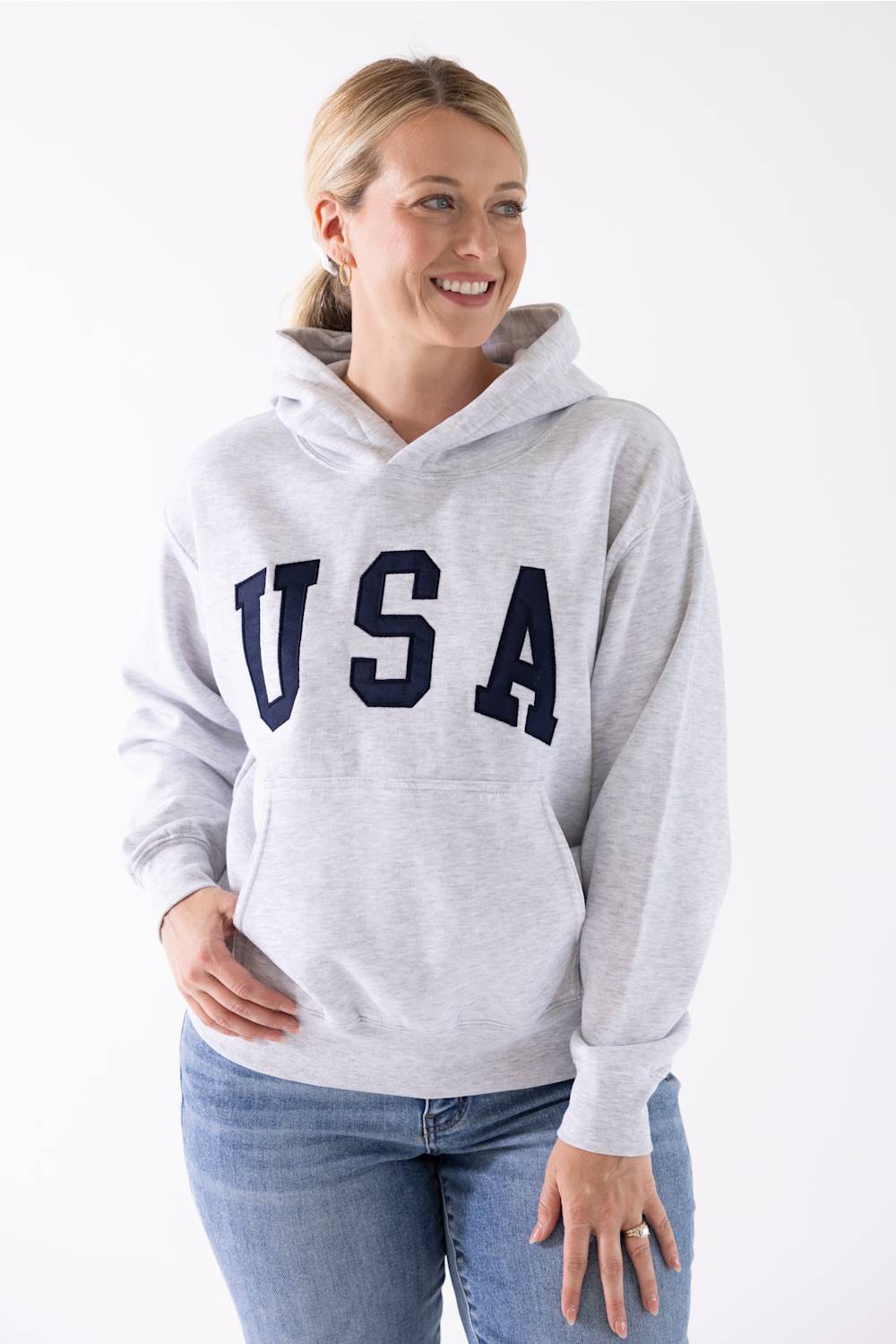Front view of a blonde woman wearing an ice grey Reflex fleece pullover hoodie with black "USA" block lettering across the chest and blue jeans.