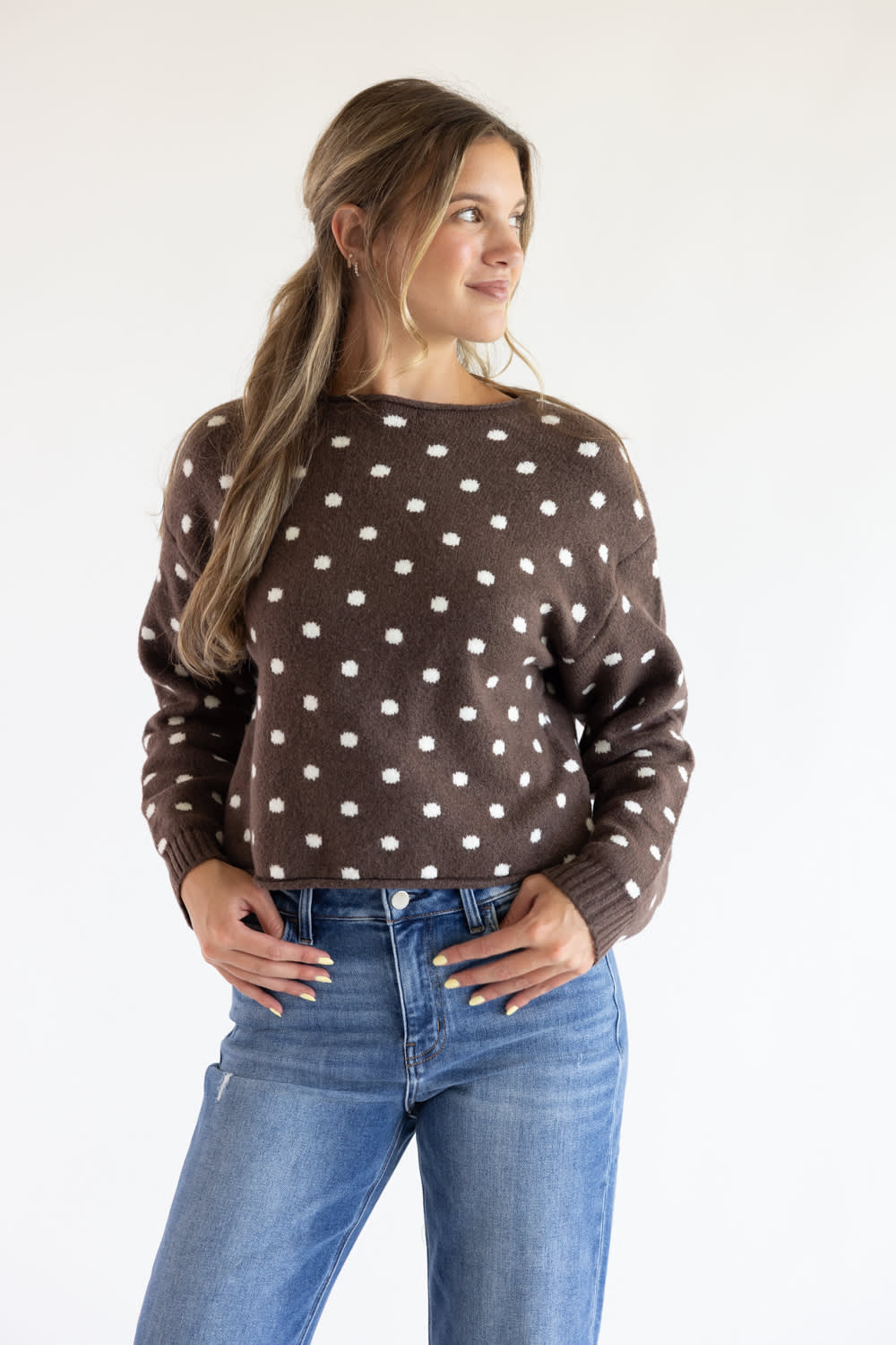 Front view of a model posing with hands in her jean pockets, wearing the Just Polly dark brown long-sleeve knit sweater featuring an ivory polka dot pattern, a slightly rolled boat neckline, and ribbed cuffs against a plain light background.