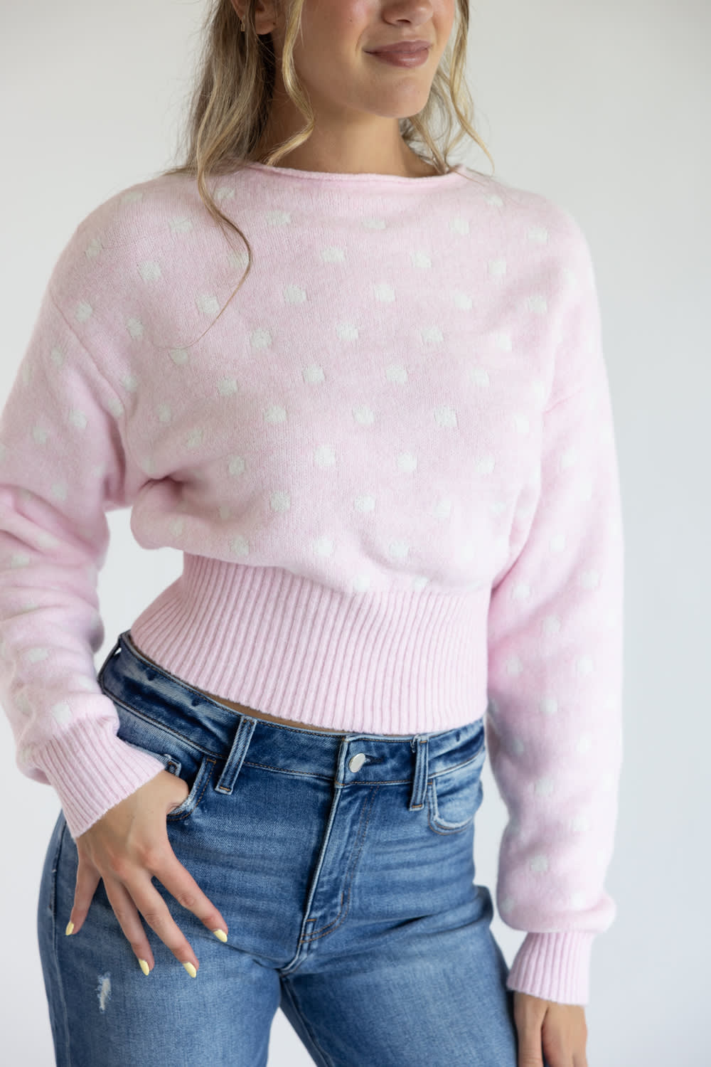 Close-up front view of a model wearing the Just Polly light pink long-sleeve knit sweater featuring a white polka dot pattern, a slightly rolled boat neckline, and a wide ribbed waistband and cuffs, styled with blue jeans against a plain white background.