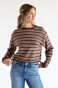 Front view of a smiling model holding the hem of her sweater, wearing the Just Polly long-sleeve knit sweater featuring horizontal stripes in brown, red, and tan, a slightly rolled boat neckline, and ribbed cuffs, styled with denim jeans against a plain white background.