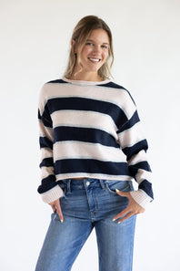 Front view of a smiling model posing with hands in her pockets, wearing the Just Polly long-sleeve knit sweater featuring wide pink and navy horizontal stripes with thin heather grey accents, styled with denim jeans against a plain background.