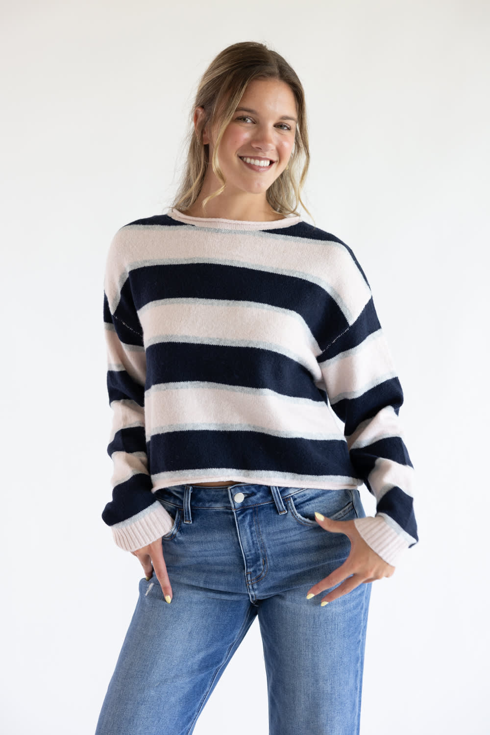 Front view of a smiling model posing with hands in her pockets, wearing the Just Polly long-sleeve knit sweater featuring wide pink and navy horizontal stripes with thin heather grey accents, styled with denim jeans against a plain background.