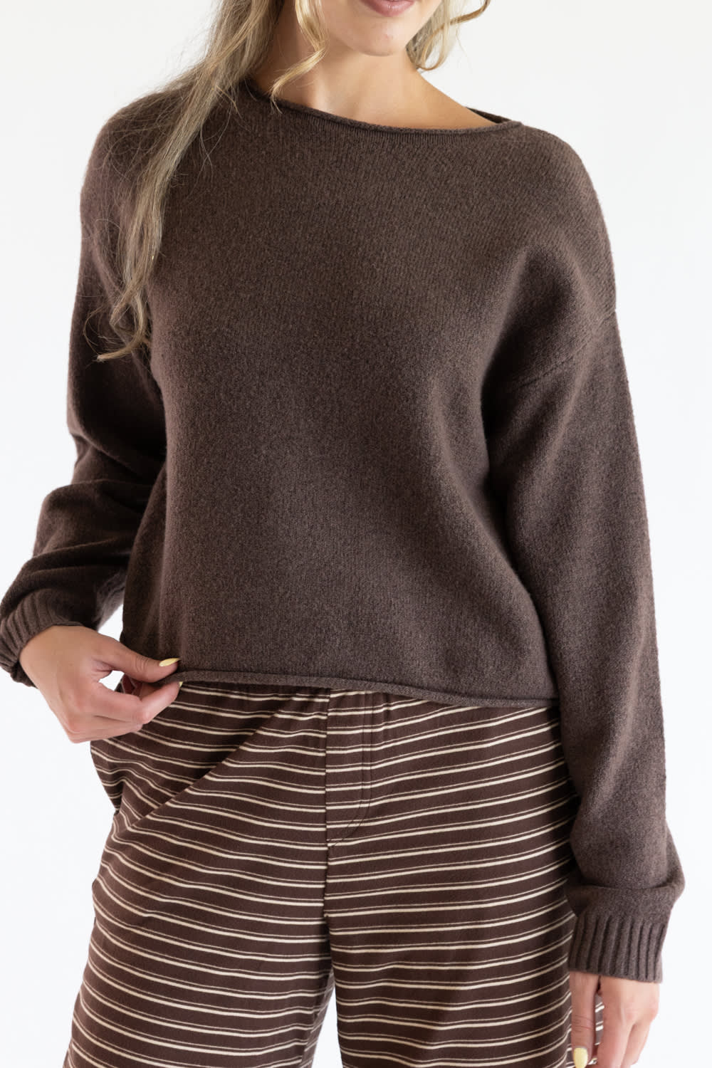 Close-up of a model wearing the Just Polly dark brown knit boat neck sweater with long sleeves, ribbed wrist cuffs, and a slightly rolled hemline, styled with brown striped pants.