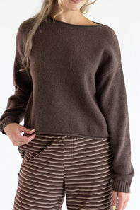 Close-up of a model wearing the Just Polly dark brown knit boat neck sweater with long sleeves, ribbed wrist cuffs, and a slightly rolled hemline, styled with brown striped pants.
