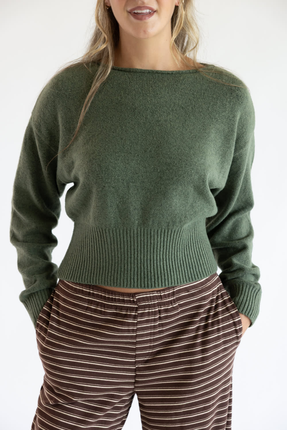 Close-up front view of a model wearing the Just Polly olive green long-sleeve knit sweater featuring a slightly rolled boat neckline and a wide ribbed waistband and cuffs, styled with brown striped pants against a plain white background.