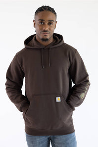 Front-facing view from the waist up of a male model against a plain white background, looking forward while wearing a dark clove brown Carhartt hooded sweatshirt with a small logo patch on the front pocket, paired with medium wash denim jeans.